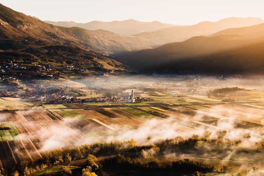 Foggy Vipava valley landscape with a village church surrounded by rolling hills and sunlit mountains.