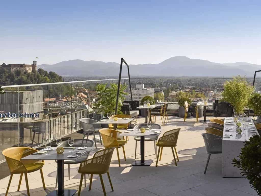 Rooftop restaurant terrace with tables overlooking Ljubljana Castle and mountains.