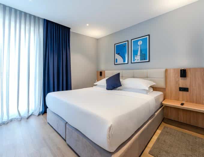 Modern hotel bedroom with large white bed, wood accents, and blue artwork near window with sheer curtains.