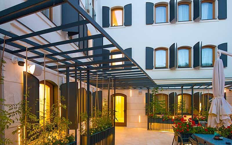 Hotel courtyard patio with metal pergola, potted red flowers, and illuminated windows.
