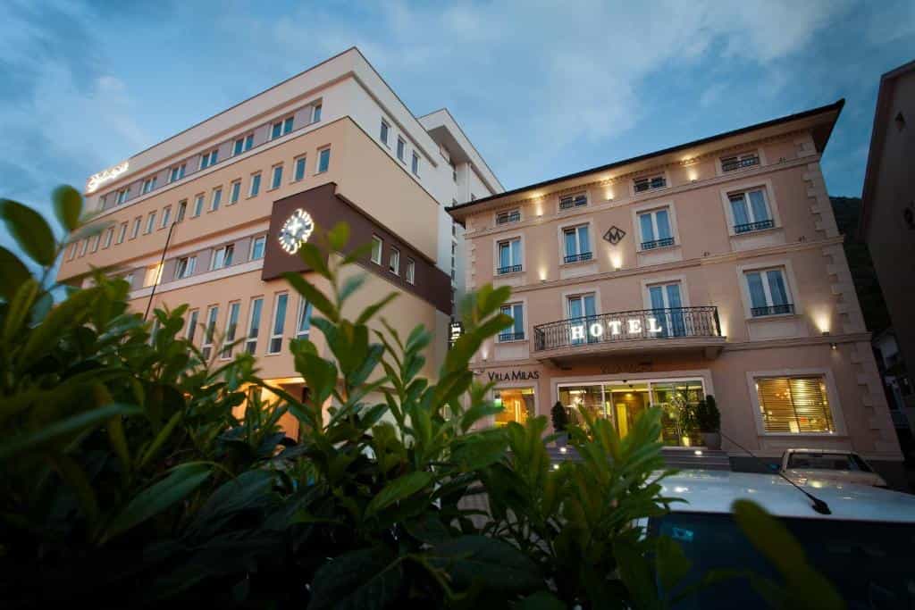 Hotel Villa Milas exterior with illuminated signage seen through dark green foliage at dusk.