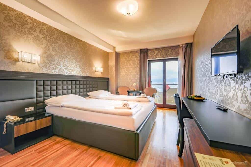 Hotel bedroom with double bed, patterned wallpaper, desk, and balcony view of water.
