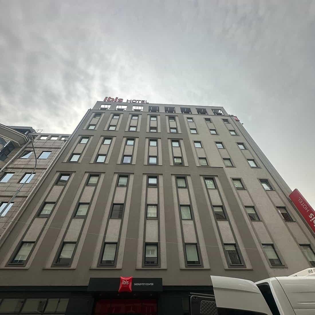 Ibis Skopje City Center hotel building facade under an overcast sky with white vans below.