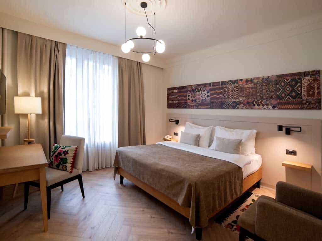 Hotel bedroom with modern light fixture, patterned wall art, and large window with sheer curtains