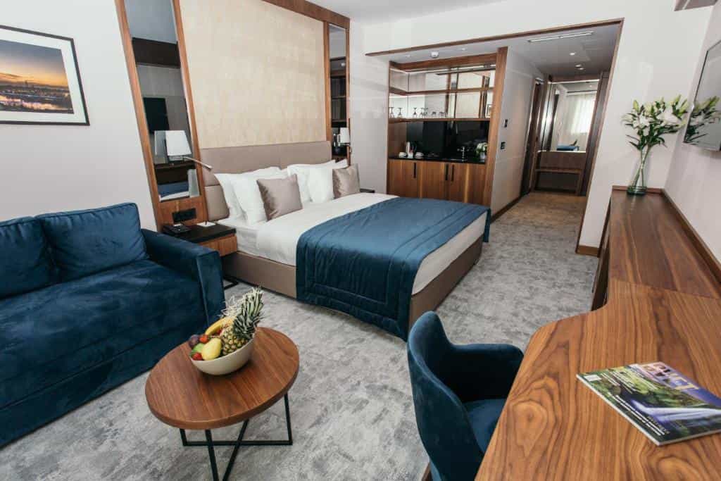 Modern hotel room with blue sofa, king bed, dark wood desk, and fruit bowl.