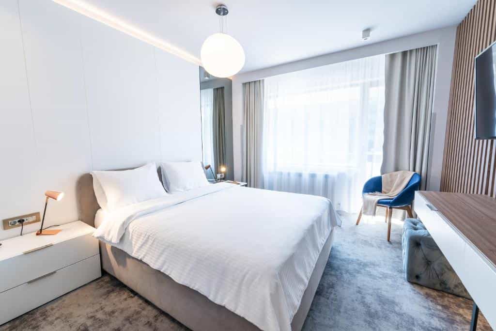 Modern hotel bedroom with white bedding, blue accent chair, and bright window light. Sinaia.