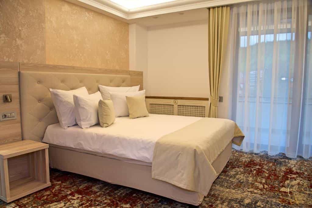 Hotel bedroom with tufted headboard, white bedding, and patterned rug near balcony doors.