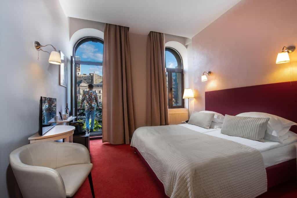 Hotel room with red carpet, large bed, and man looking out arched balcony window at city.