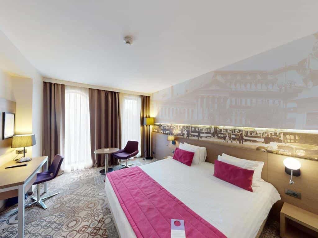 Hotel room with white bedding, pink accents, desk, and city mural above the bed.