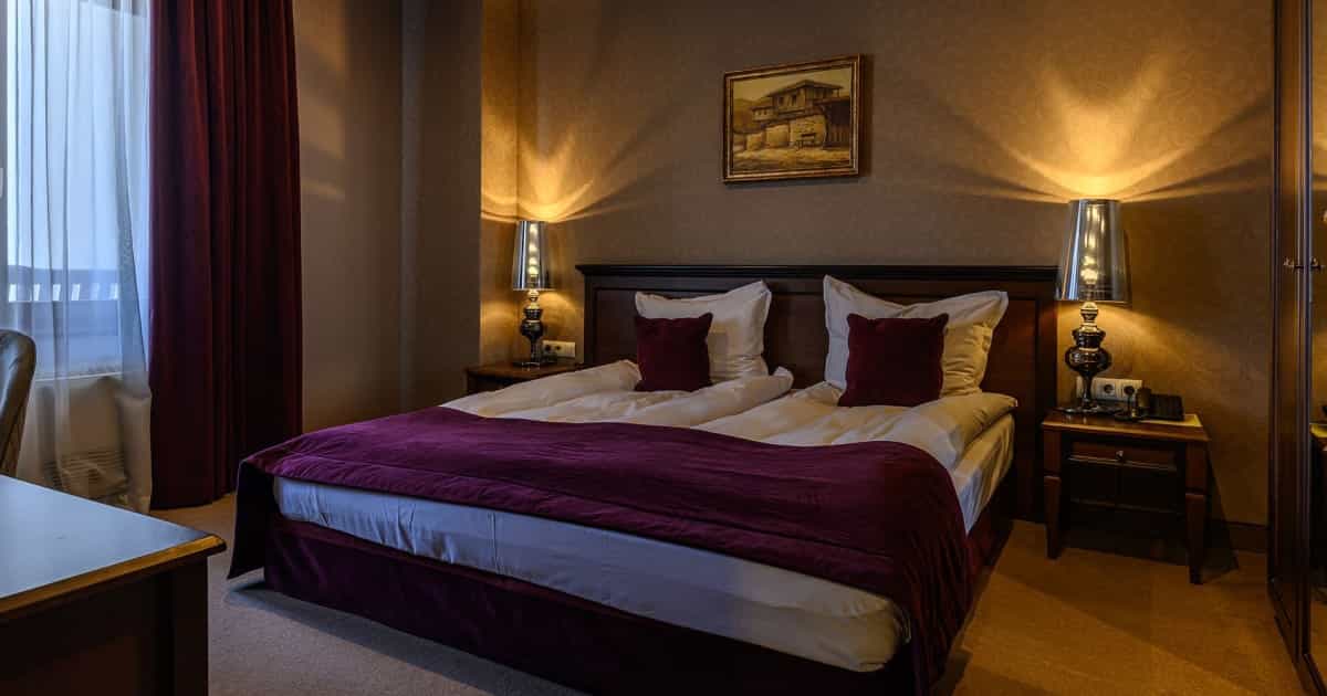 Hotel bedroom with twin beds, purple bedding, and illuminated bedside lamps near a window with dark curtains.