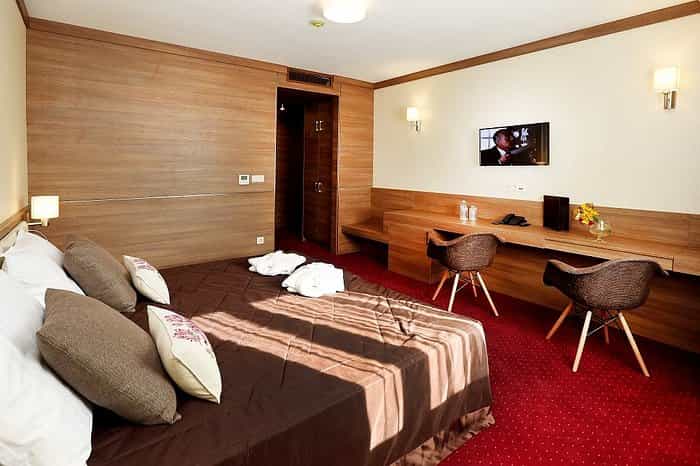 Hotel room with brown bedding, red carpet, wood paneling, and two wicker chairs at a desk.