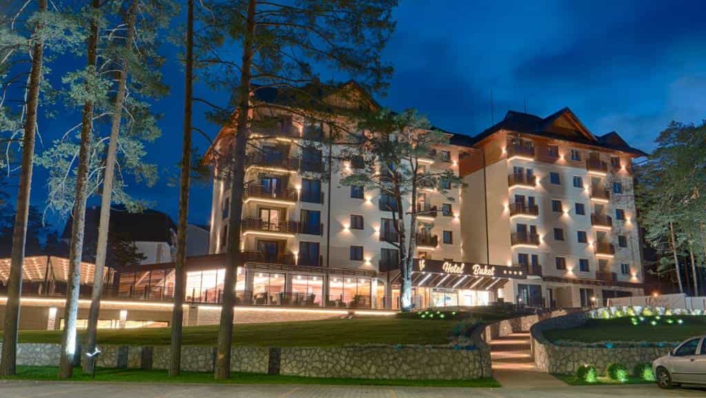 Hotel Buket illuminated at night, surrounded by tall pine trees in a mountainous setting.