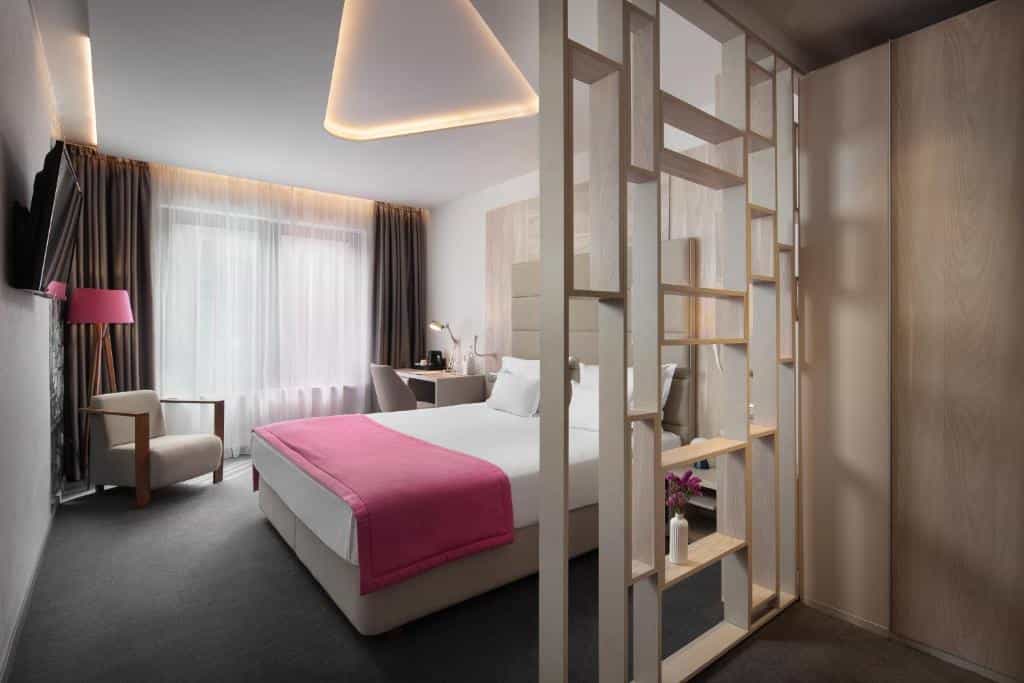 Modern hotel room with white bedding, pink throw, armchair, desk, and wooden shelving divider in Colors Inn Sarajevo.