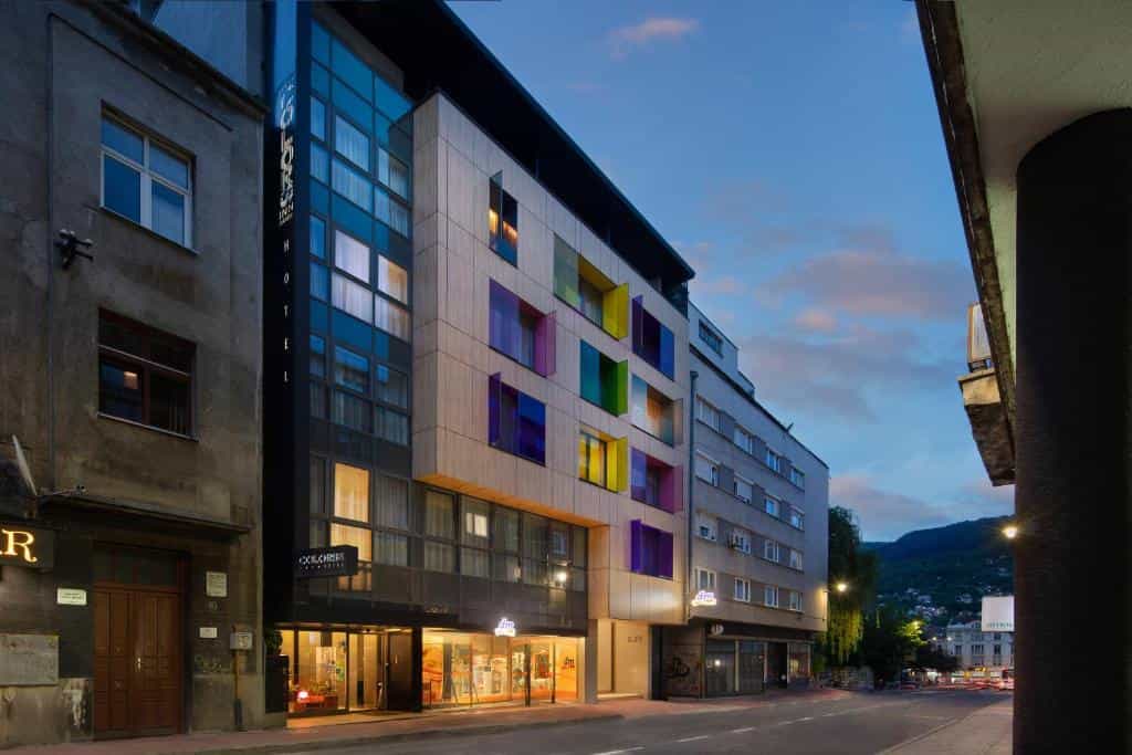 Colors Inn Sarajevo hotel exterior with colorful window panels at dusk, mountains in background.