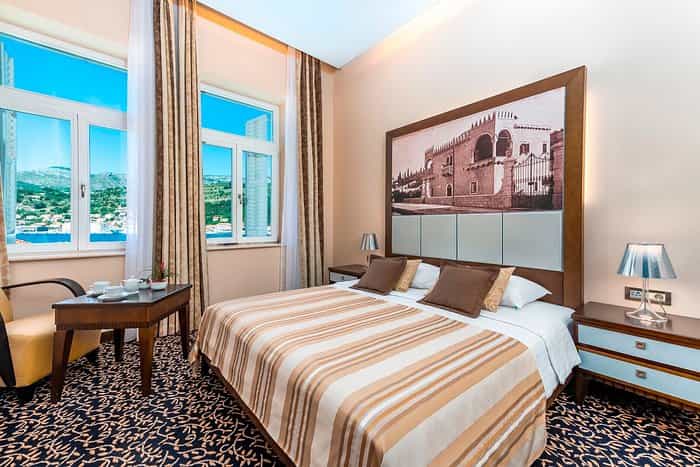 Hotel room with striped bedspread, view of coastal town, and historic artwork above headboard