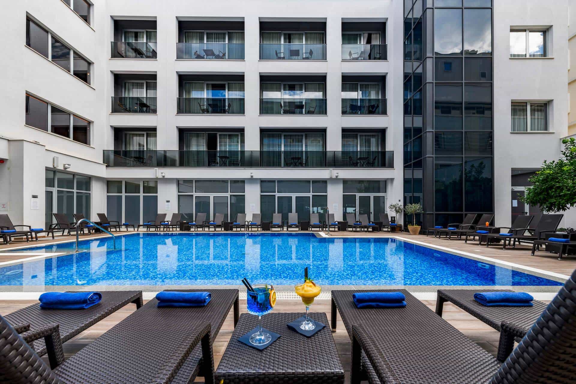 Resort swimming pool with lounge chairs, blue cocktails, and modern white hotel building.