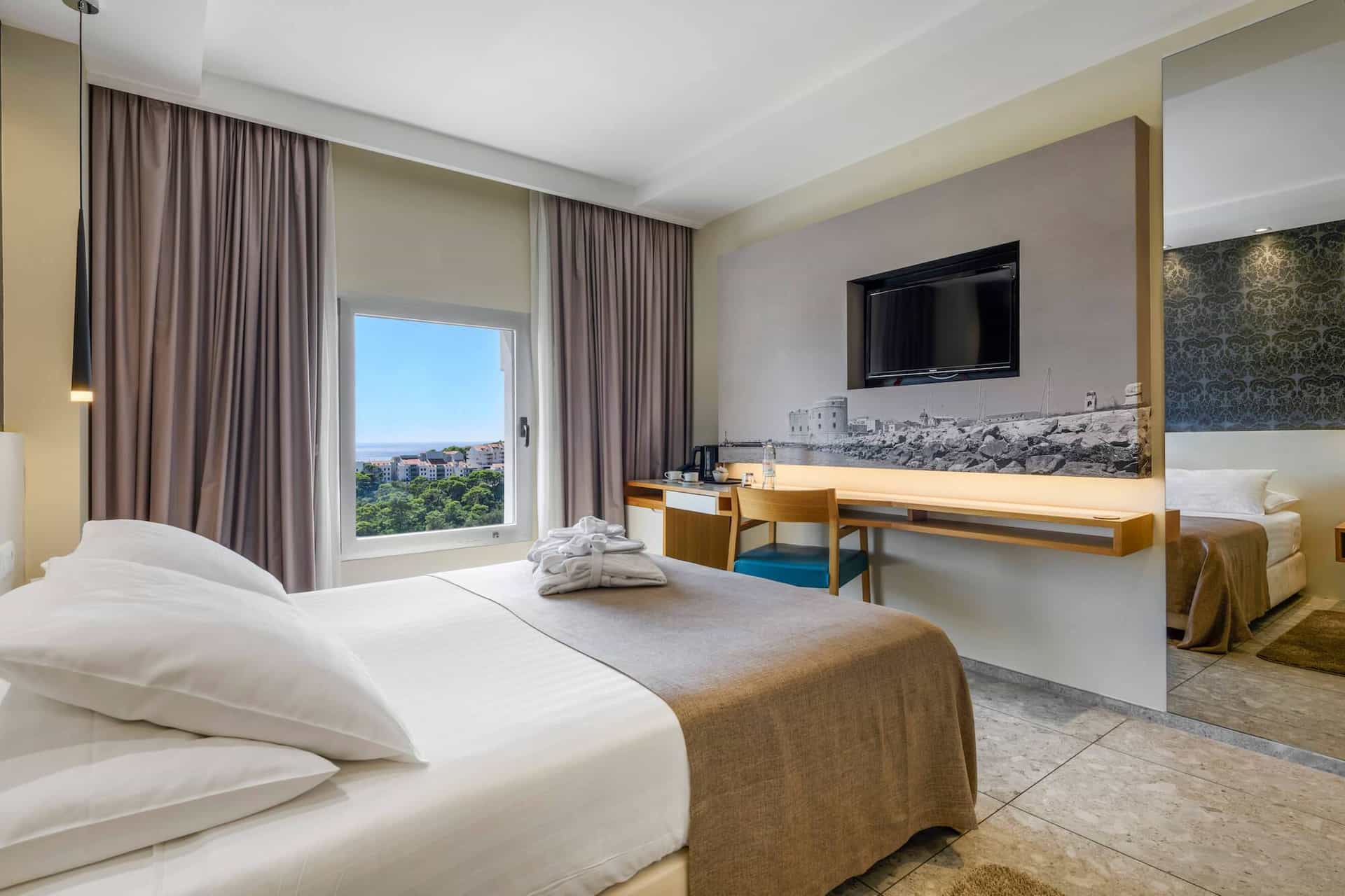 Hotel room with made bed, desk, and window view of coastal town and sea