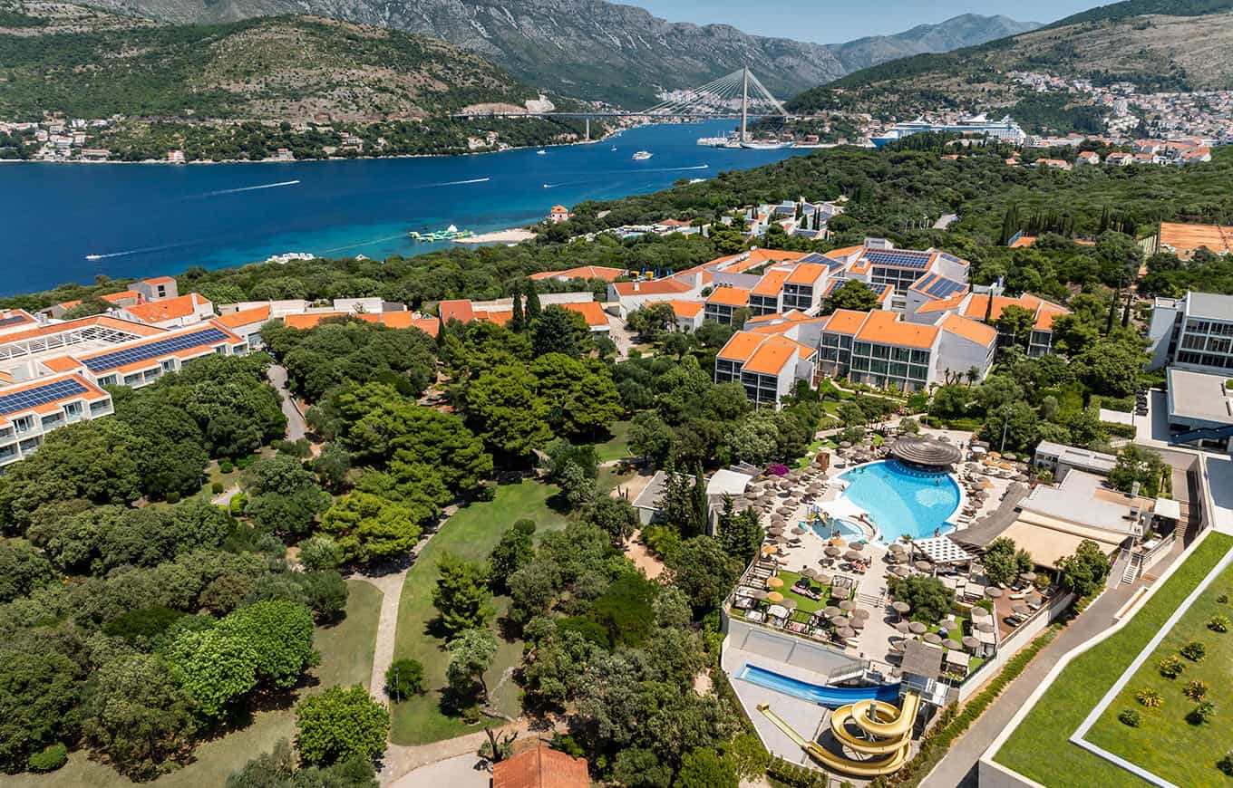 Resort with orange-roofed buildings, pool, and water slide overlooking the sea and bridge in Dubrovnik.
