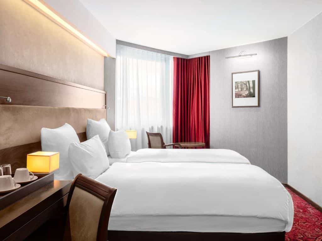 Twin beds with white linens, bedside lamps, and red curtains in a Movenpick hotel room in Sarajevo.