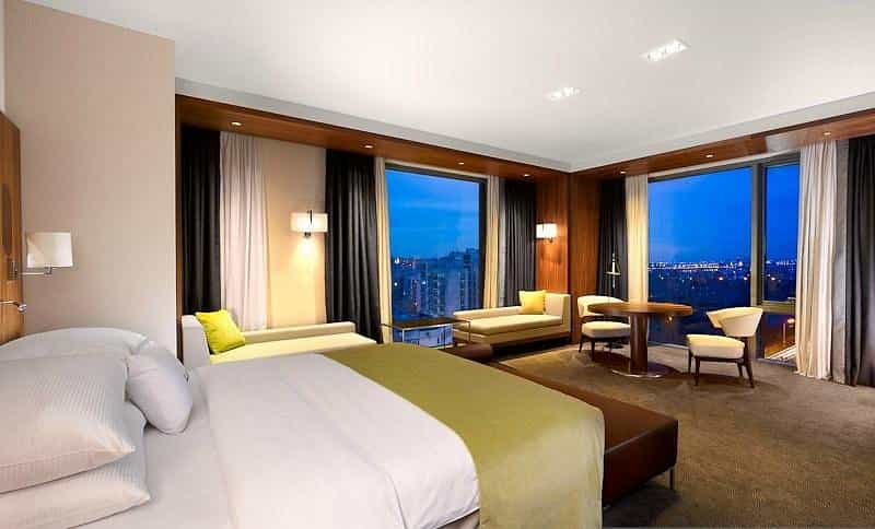 Modern hotel room with large windows showing a city skyline at dusk.