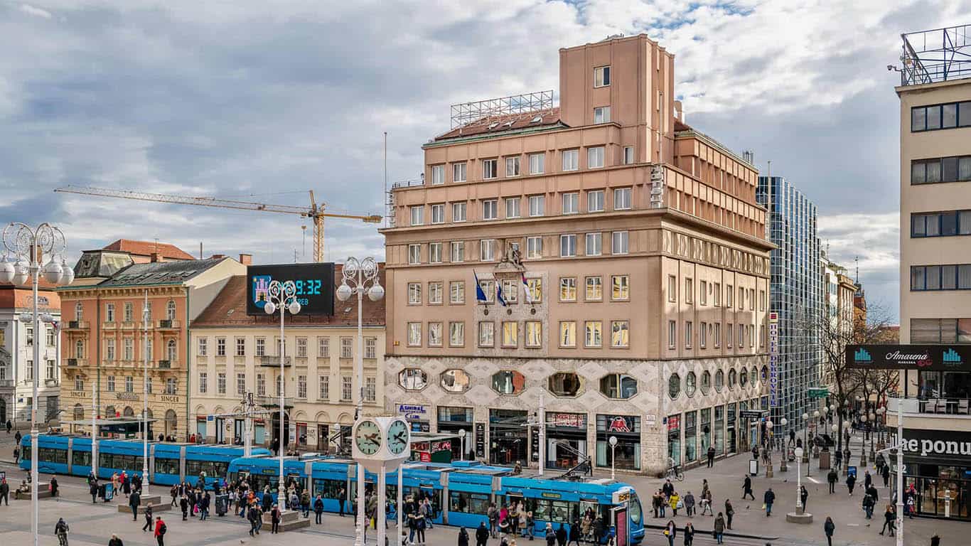 Blue trams and pedestrians in a busy city square with historic buildings, Zagreb.