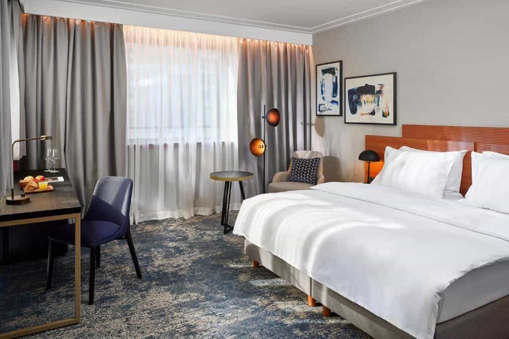 Hotel bedroom with large white bed, dark desk, blue chair, and sheer curtains over window.