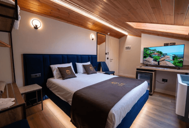Hotel room with blue bed, wood ceiling, and television displaying a hillside town view, Krujë.
