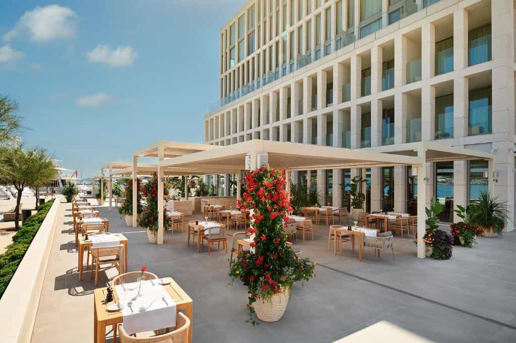 Outdoor dining terrace with tables under pergolas next to a modern hotel and marina.