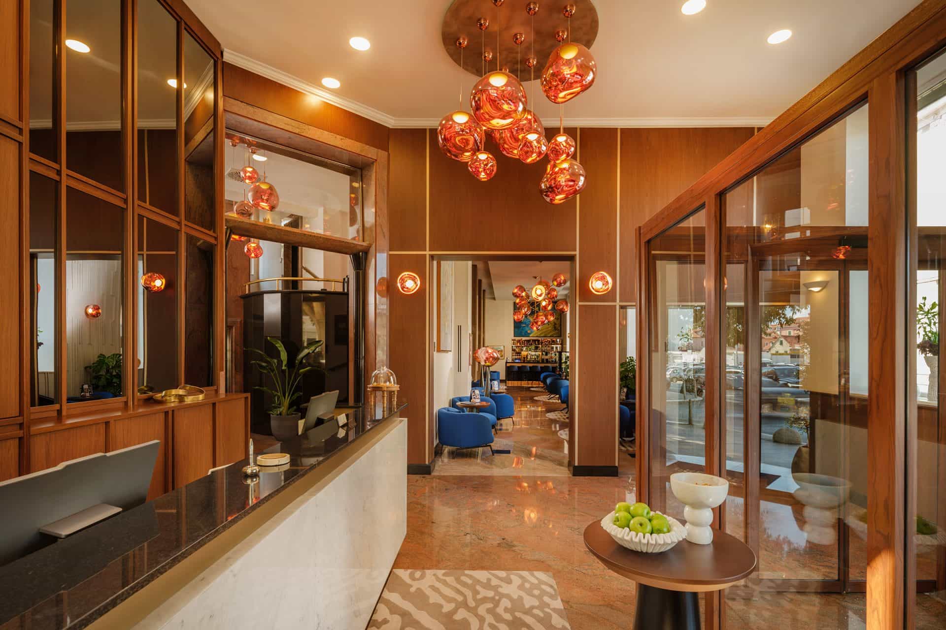 Hotel reception area with wood paneling, marble counter, and modern orange glass chandeliers.