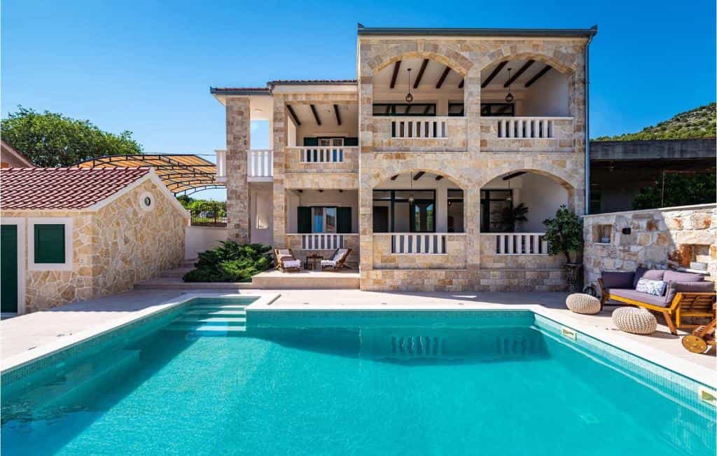 Stone villa with balconies overlooking a bright blue swimming pool on a sunny day.