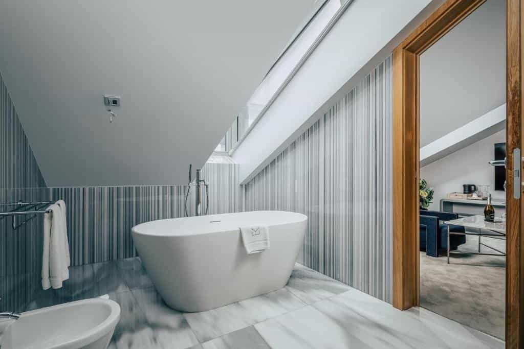 Modern bathroom with freestanding white bathtub, marble floor, and skylight, view into hotel room.
