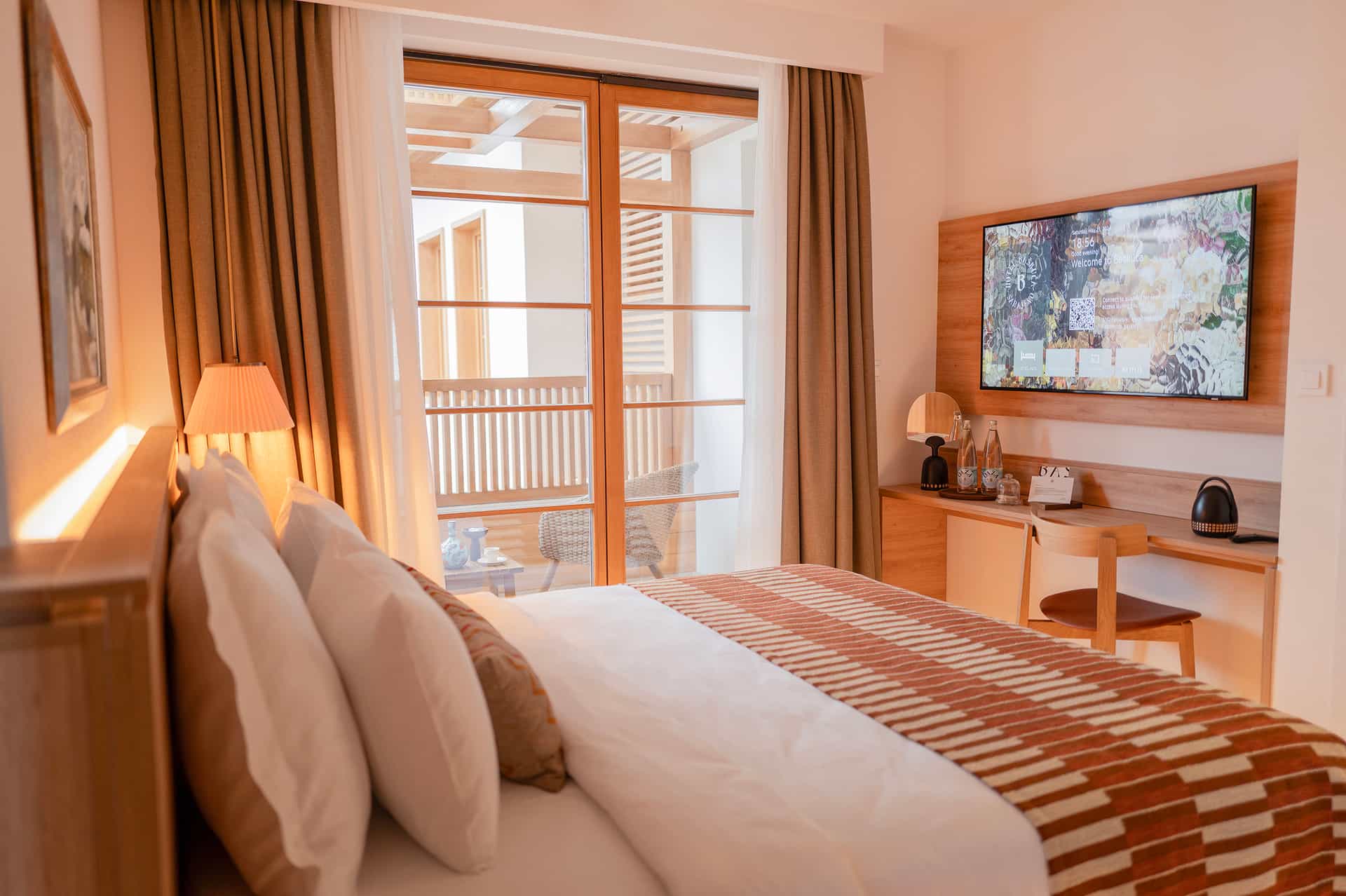 Hotel room interior with large bed, TV displaying Hotel Baškuca Sarajevo welcome screen, and balcony access.