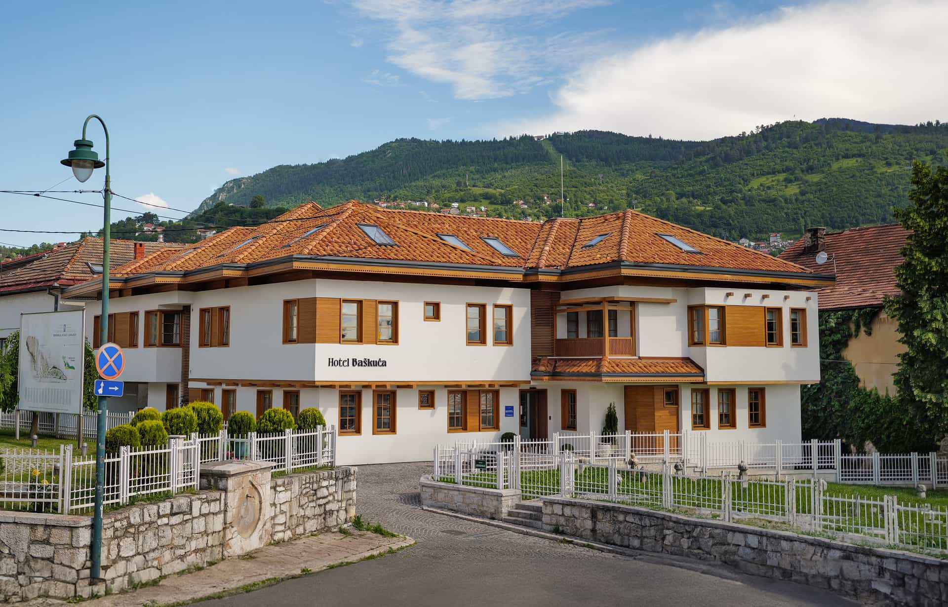 Hotel Baščuća with white facade and terracotta roof against green forested mountains.