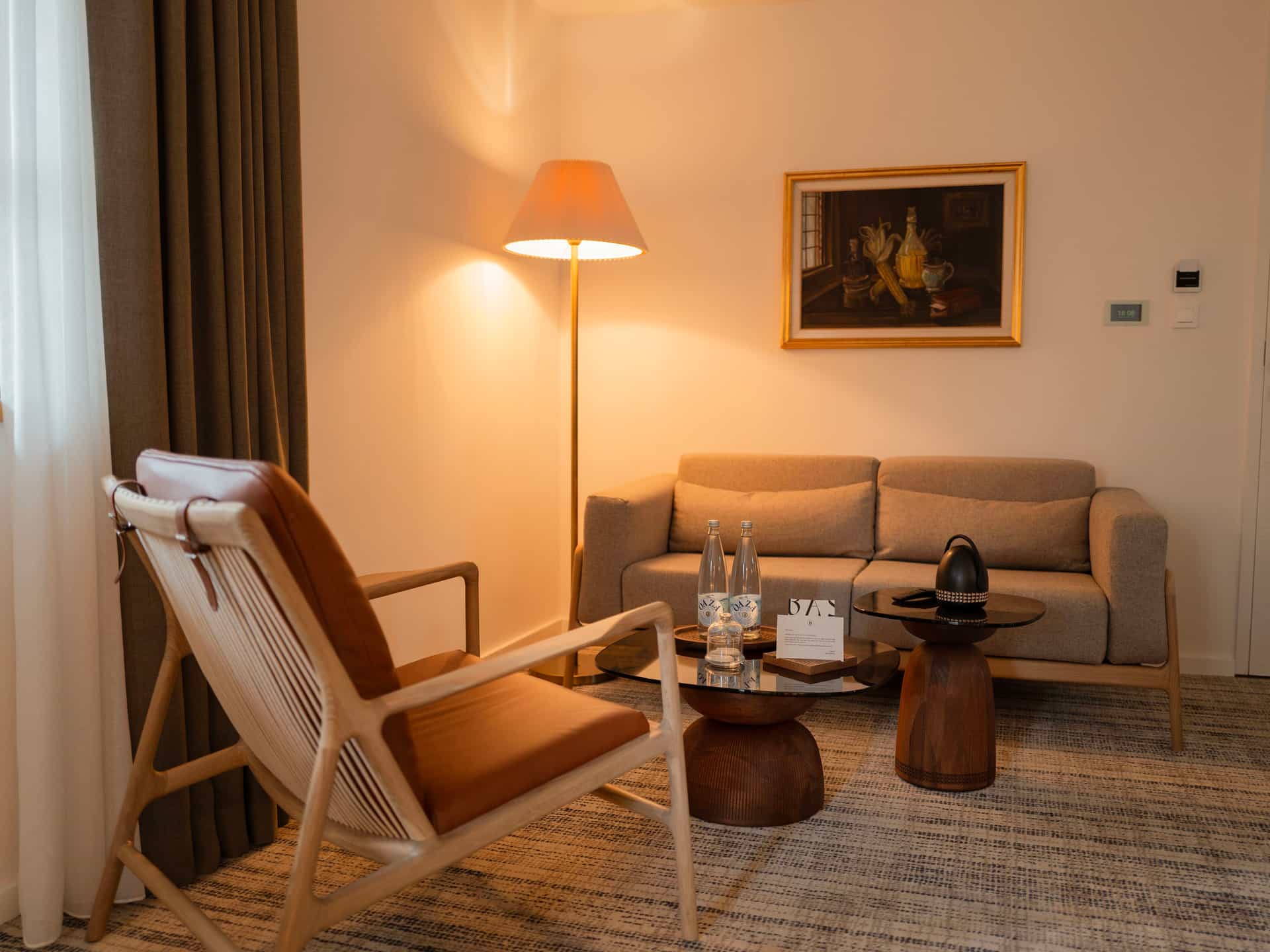 Hotel sitting area with modern wood chair, sofa, floor lamp, and still life painting.