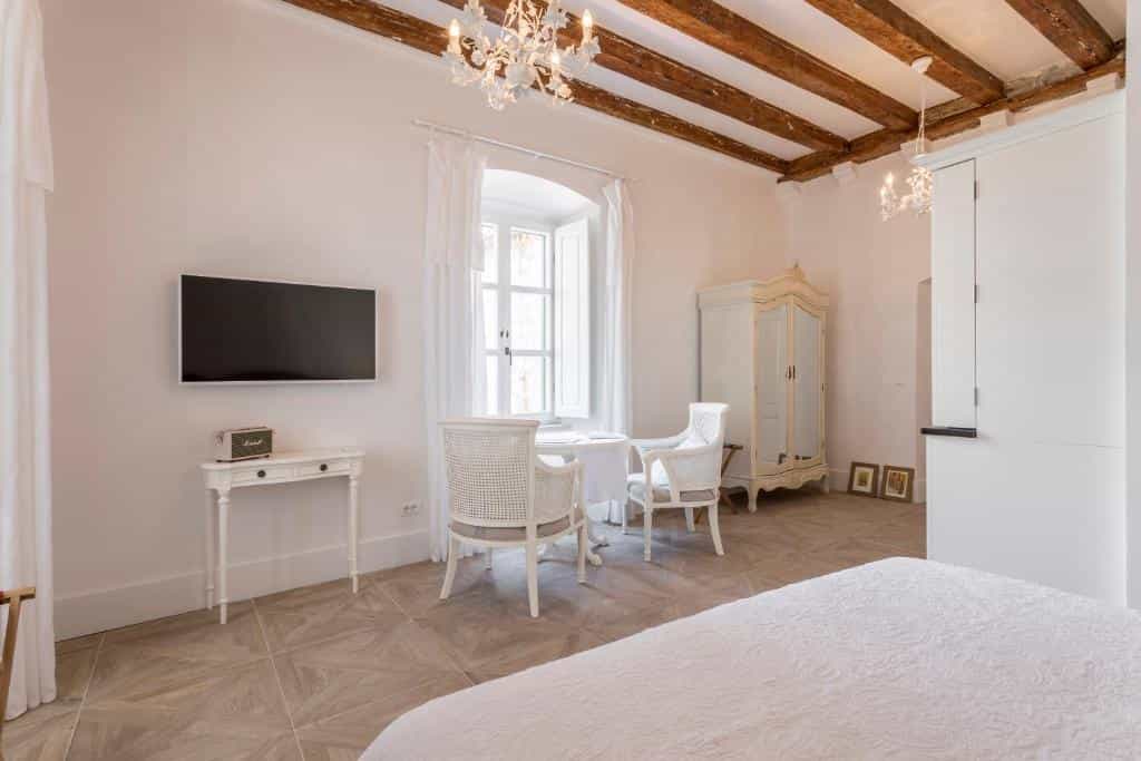 Bright hotel room with white furniture, exposed wooden ceiling beams, and a view through a window.