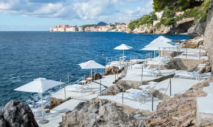 Sun loungers and white umbrellas on tiered concrete terraces overlooking the sea near Dubrovnik city walls.