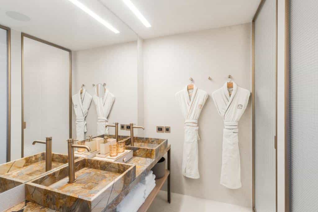 Hotel bathroom with dual marble sinks, bronze fixtures, and two white bathrobes hanging on the wall.