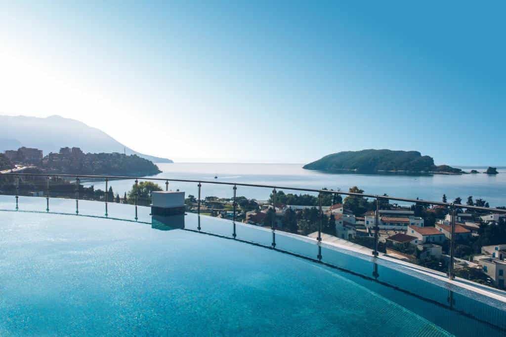 Infinity pool overlooking coastal town, sea, and forested island under clear blue sky.