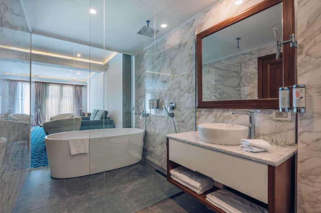 Modern hotel bathroom with freestanding tub, marble walls, and view into adjoining living area.