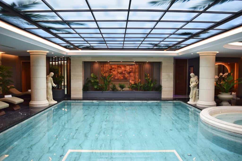 Indoor swimming pool with classical statues, plants, and a skylight showing palm fronds, Xheko Imperial Hotel Tirana.
