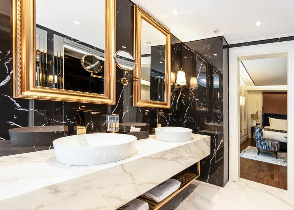 Luxury bathroom with dual white vessel sinks, gold mirrors, and black marble walls, opening to a bedroom.