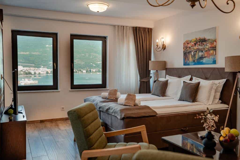 Hotel room with bed and green chair overlooking water and green mountains through windows.