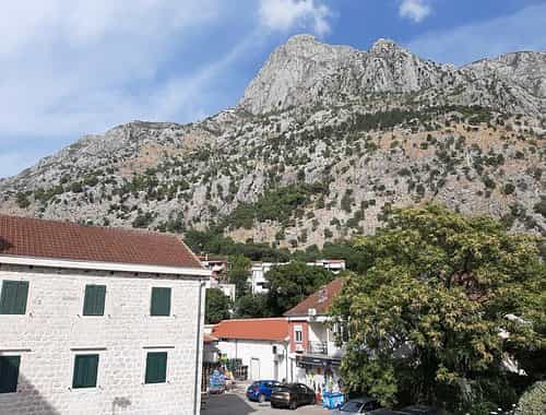 Stone building with green shutters below a large, scrub-covered mountain under a blue sky.