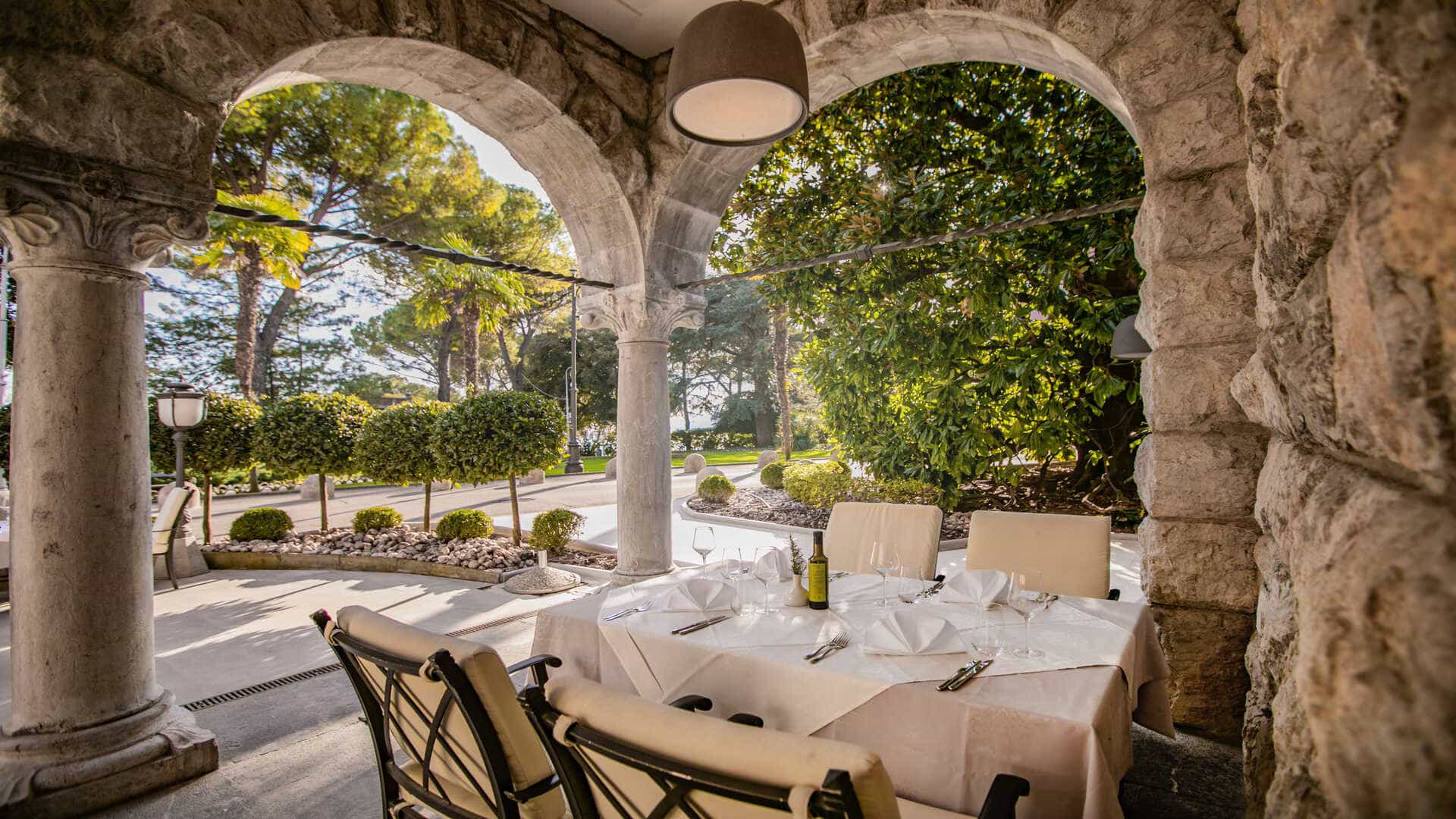 Outdoor dining table set under stone arches overlooking a sunny, tree-lined coastal area.