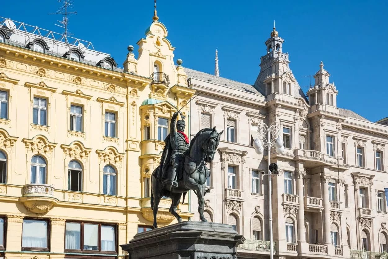 Equestrian statue in Zagreb main square before ornate yellow and white historic buildings.