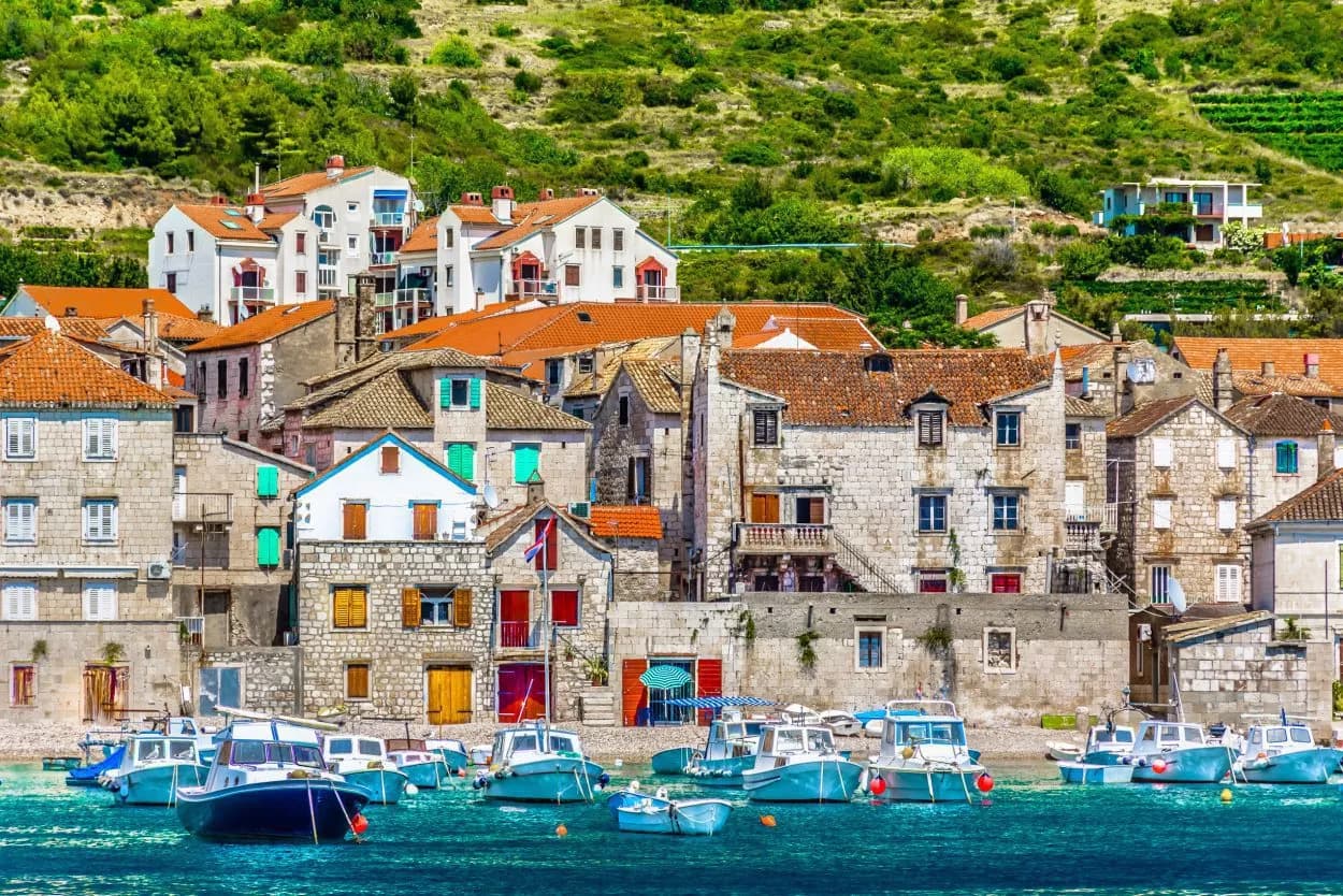 Boats moored in turquoise water near stone houses with terracotta roofs in Komiza village on Vis.
