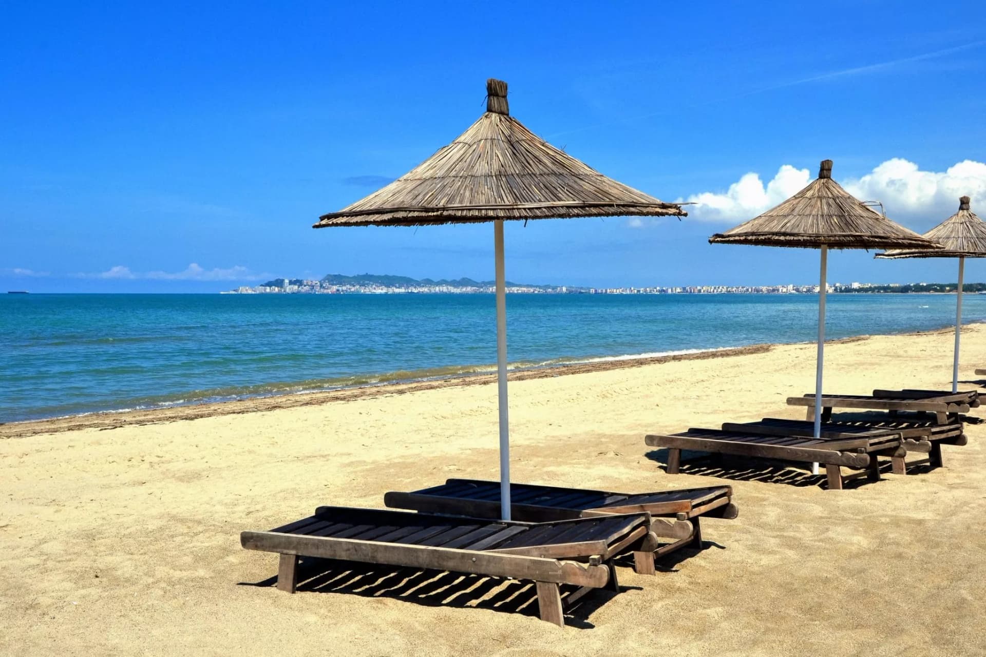 Beach with straw umbrellas and wooden loungers facing the sea near Durres city skyline.