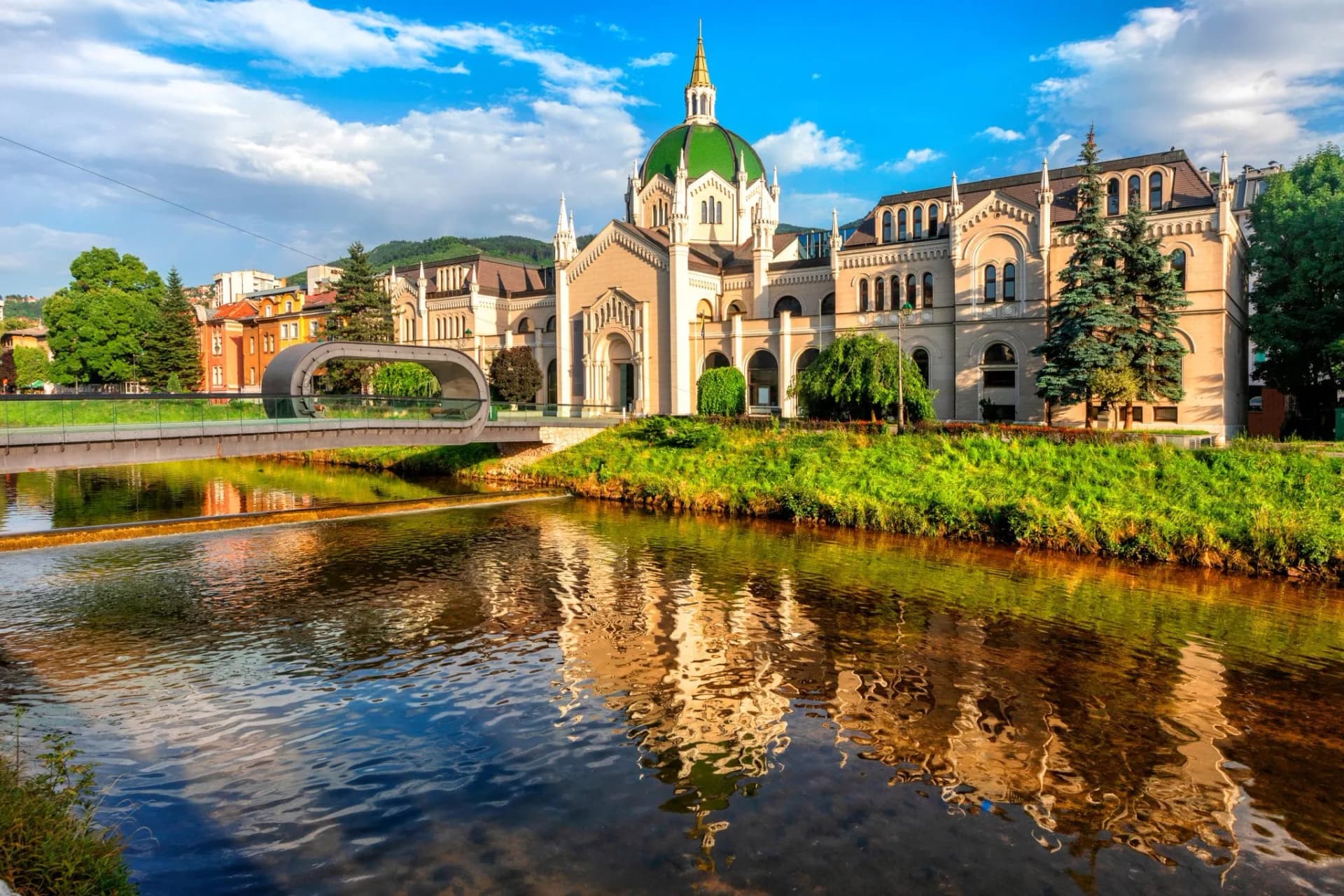Sarajevo City Center building with green dome reflected in river near modern bridge.