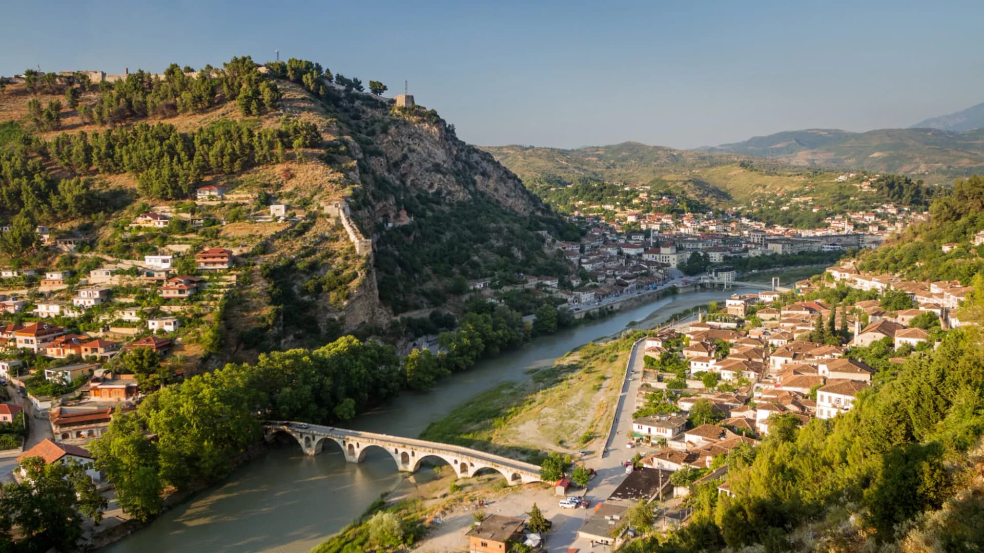 Panorama of Berat, Albania, showing houses climbing a hill above a river and stone arch bridge.