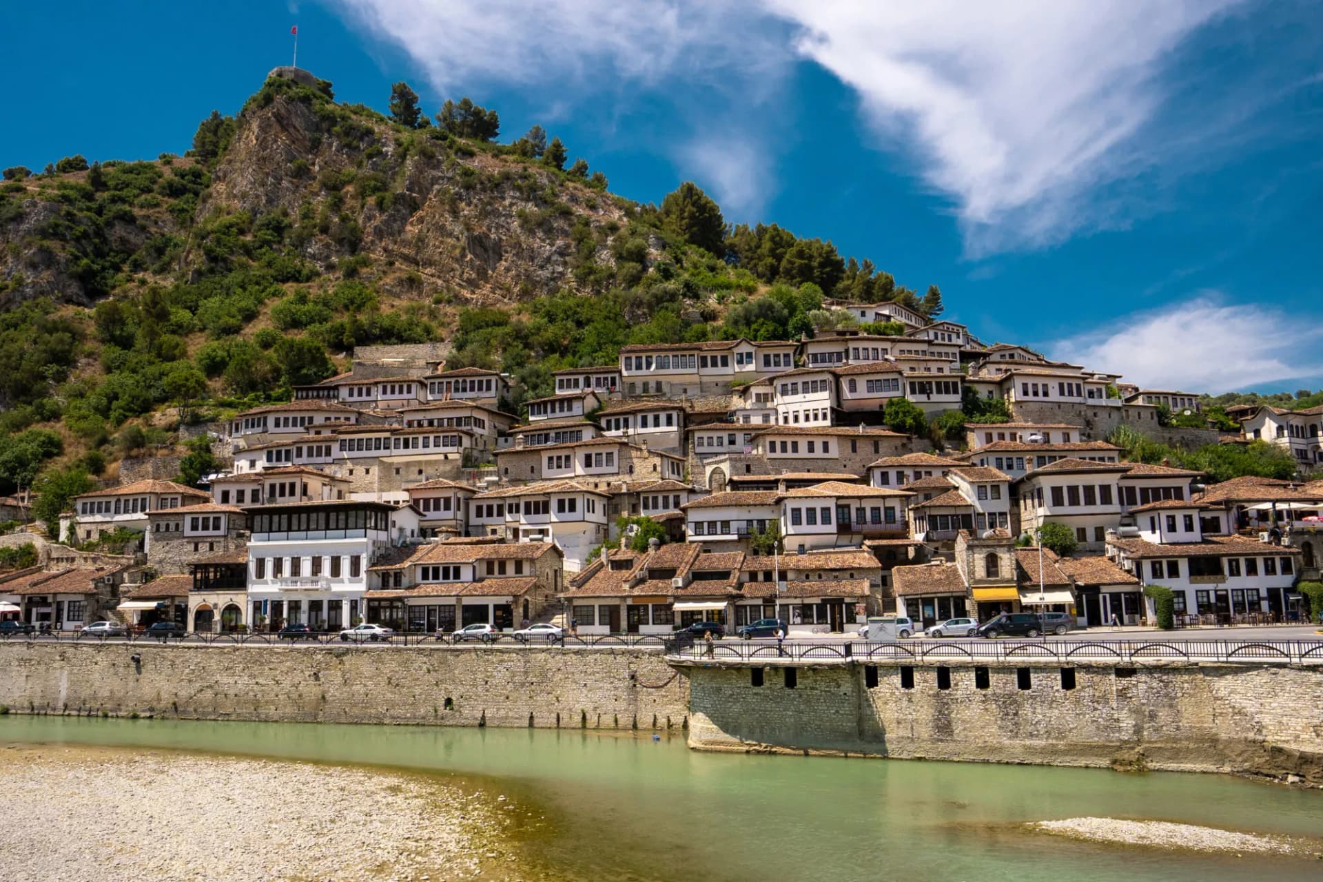White Ottoman-style houses cascade up a hill above a river, with a fortress on the peak.